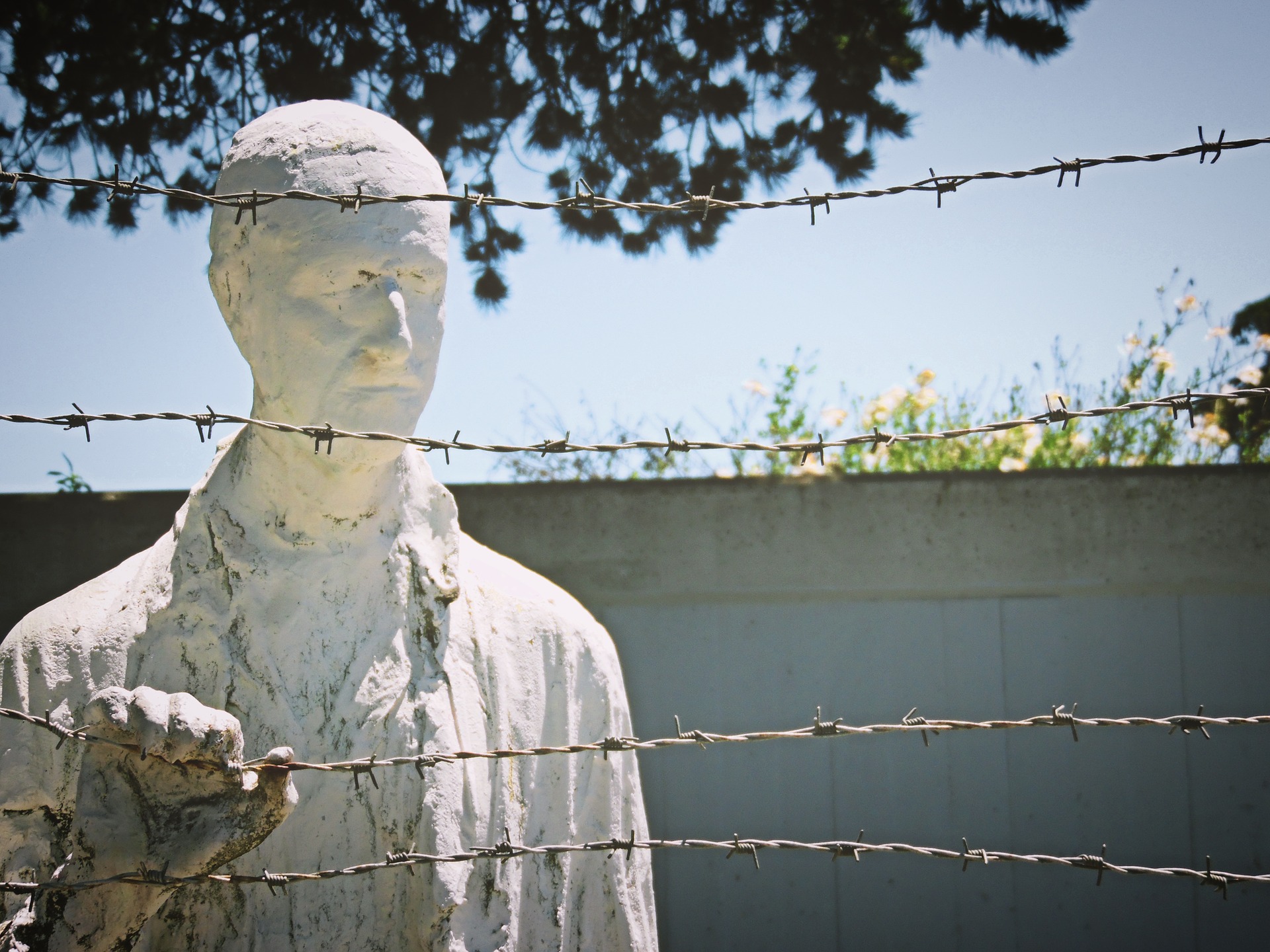 A Holocaust memorial. A statue of a man on the left behind barbed wire. His right hand rests on the barbed wire. A concrete wall in the background.