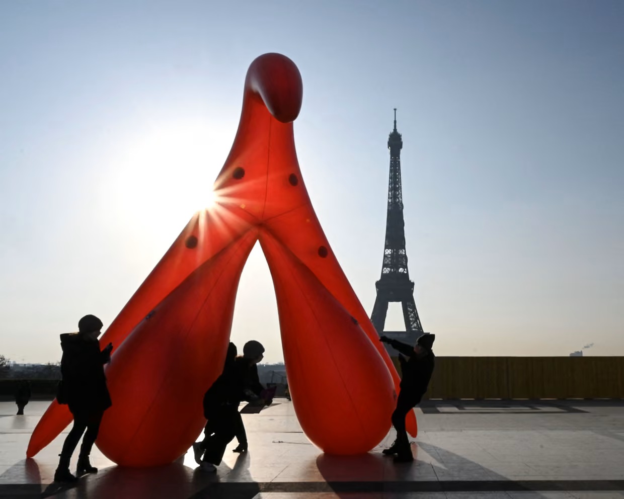 Members of the group Gang du Clito demonstrate against sexual illiteracy in 2021 Paris. Against the backdrop of the Eiffel Tower, a giant model clitoris in red, about 4 to 5 metres tall, is positioned by a group of four humans. Due to the perspective, the clitoris appears as tall as the tower.