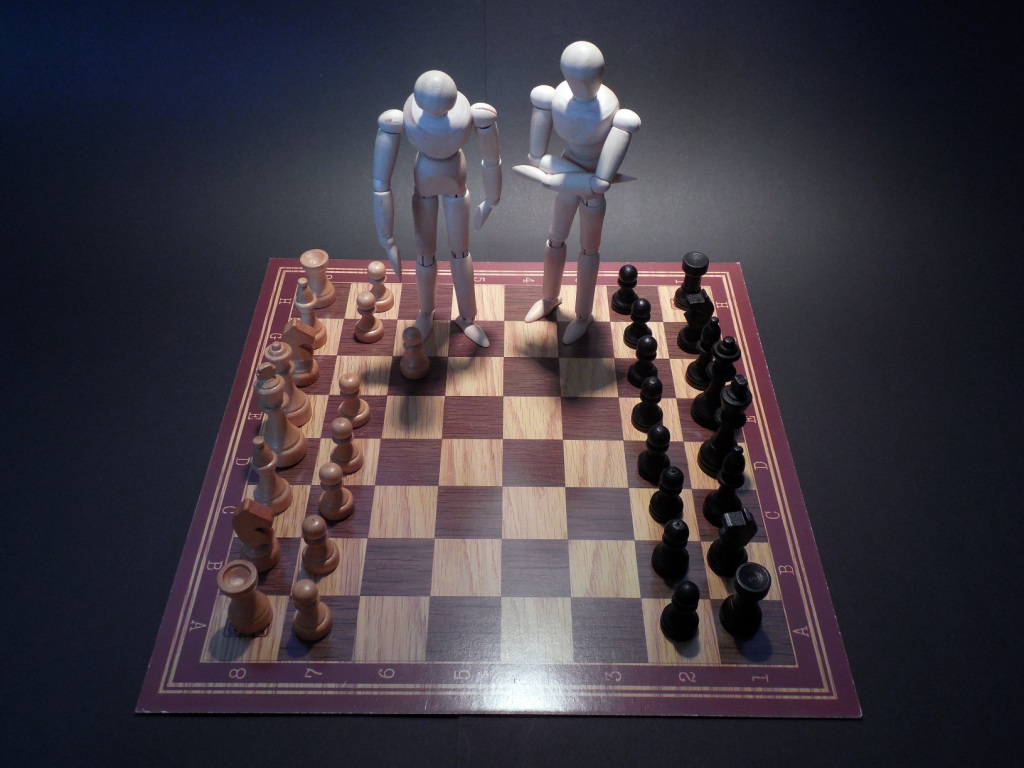 A chess board set up with all its pieces, but two additional giant white figures walk over the board, intended to give the impression of white supremacy