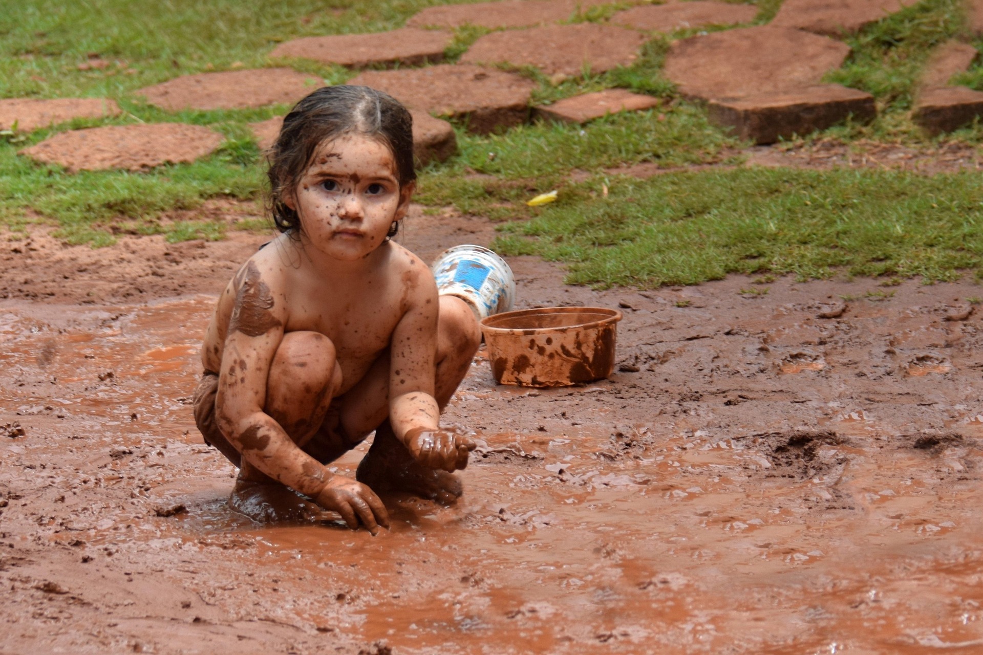It is a sunny day. A young dark-haired and dark-skinned girl is crouched down in a huge mud puddle. She plays with the mud. Nearby are her containers for play. Behind her is grass and a path.