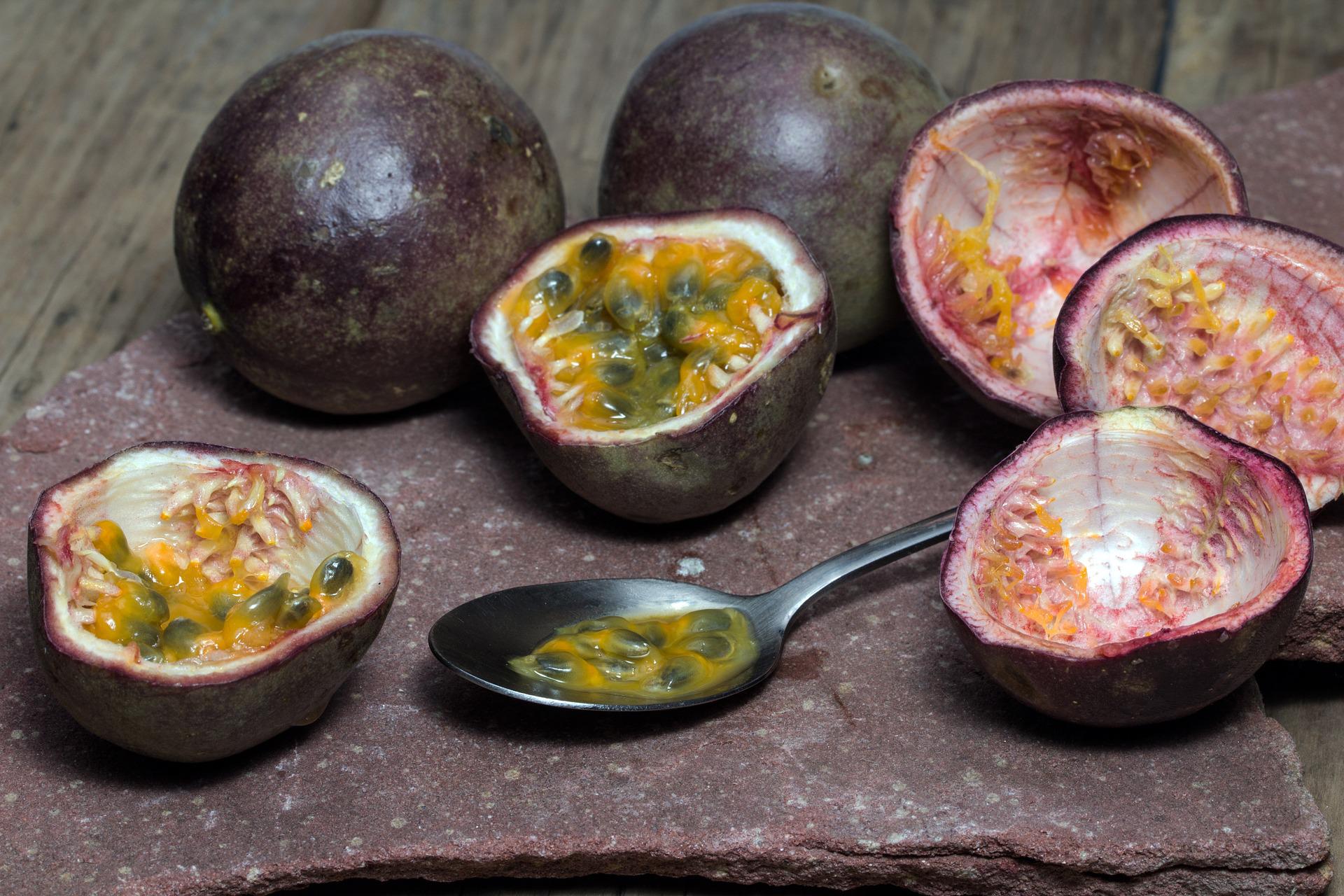 On a stone plate on an old wooden table are some passion fruit. Two remain unopened. The rest have been halved, some eaten with a small spoon.