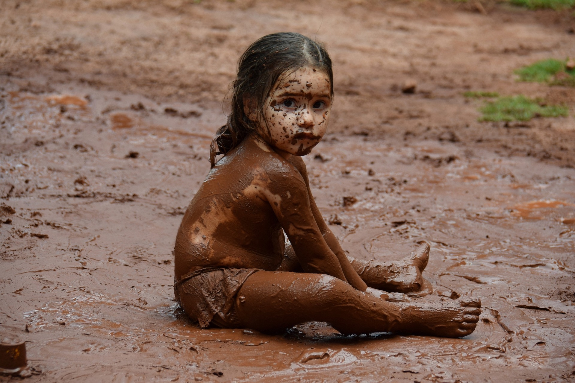 It is a sunny day. A young dark-haired and dark-skinned girl is sitting in the middle of a huge mud puddle. Her whole body is covered in mud! She looks like she has been enjoying herself.
