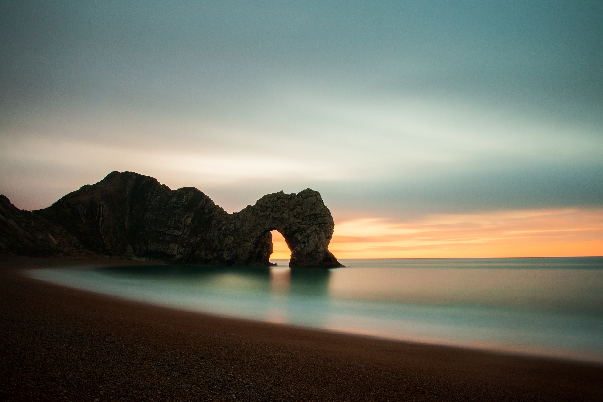 At the darkened beach, we gaze at Durdle Door (a giant natural arched rock formation in Dorset, England). Dawn is imminent, the horizon is lighting up. The sky is a beautiful blue. The photo is long-exposure, so the sea's movements are slightly blurred.