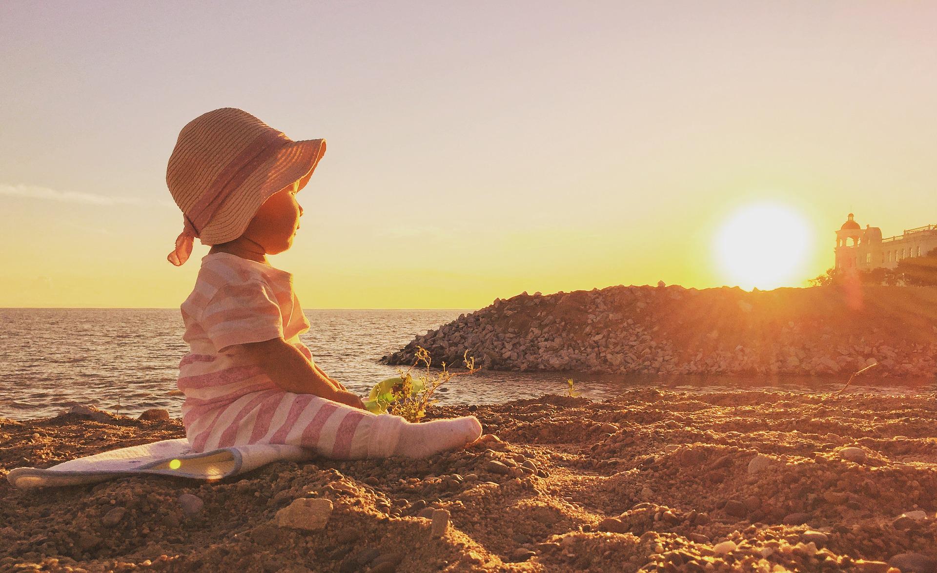 A serene toddler in a white and pink striped onesie and straw hat is seated on a sandy beach. He/she is gazing at the sunrise. The sun is rising over a stony promontory and its village.