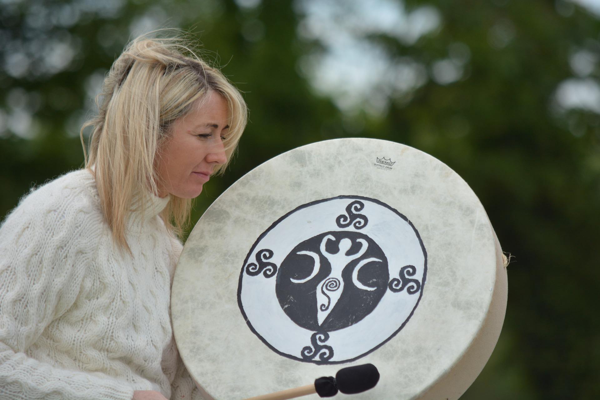 A woman with blonde hair and white jumper is drumming. She is moving. It seems just before sunrise. Her drum has a Goddess image at its centre.