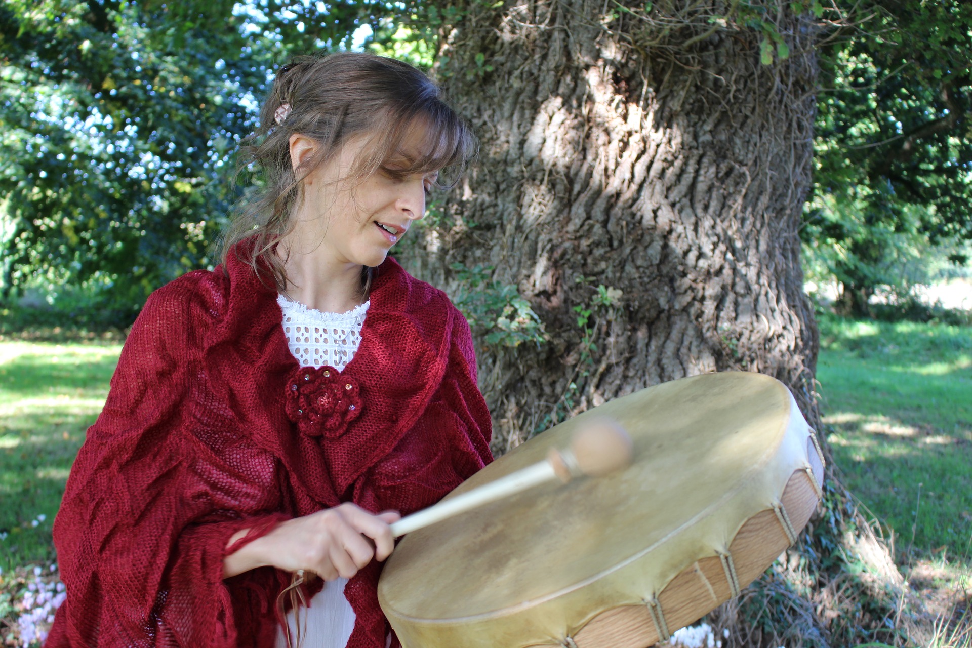 A woman in a druidic red cape is drumming. She is standing in front of an oak tree. The sun is up now.