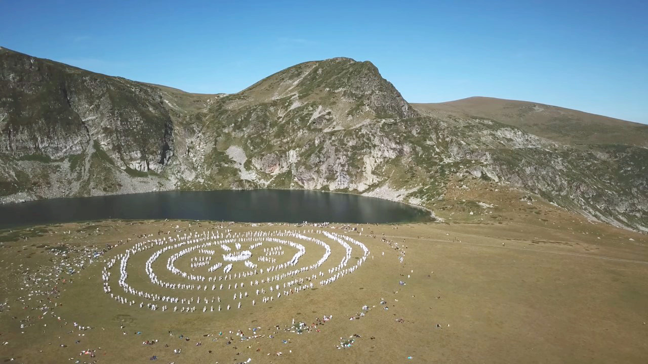 High in the Rila Mountains of Bulgaria, by a lake, in the morning sun, we see a large group of people dressed in white. They are practising Paneurhythmy, specifically the part called 'Sun's Rays'. At the centre is a circle of people and rays of people branch out from the circle, so it looks like a sun symbol. Further circles of dancers surround the sun symbol.