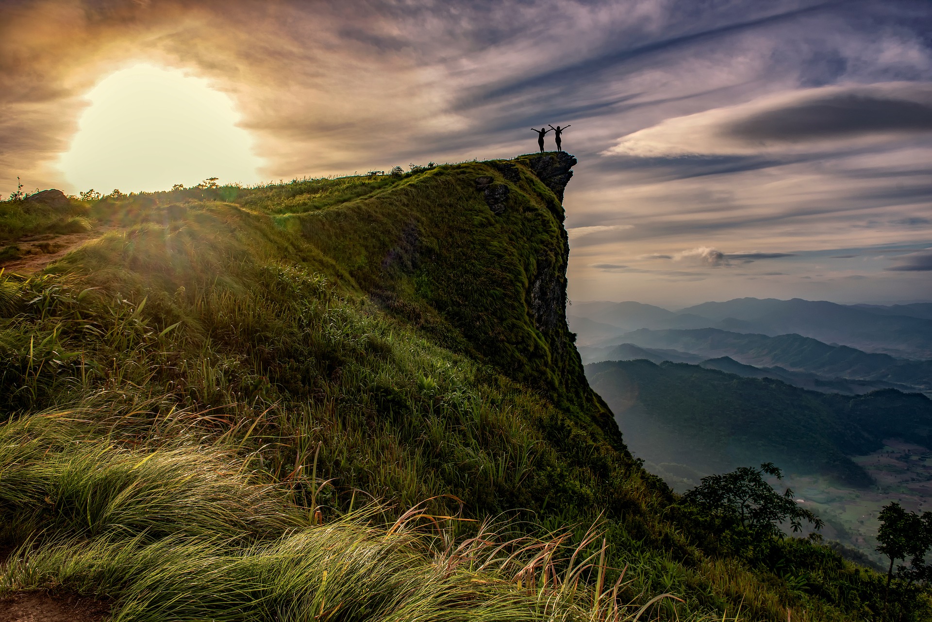 Atop a grassy cliff, in the distance two women raise their hands and greet the dawn sun