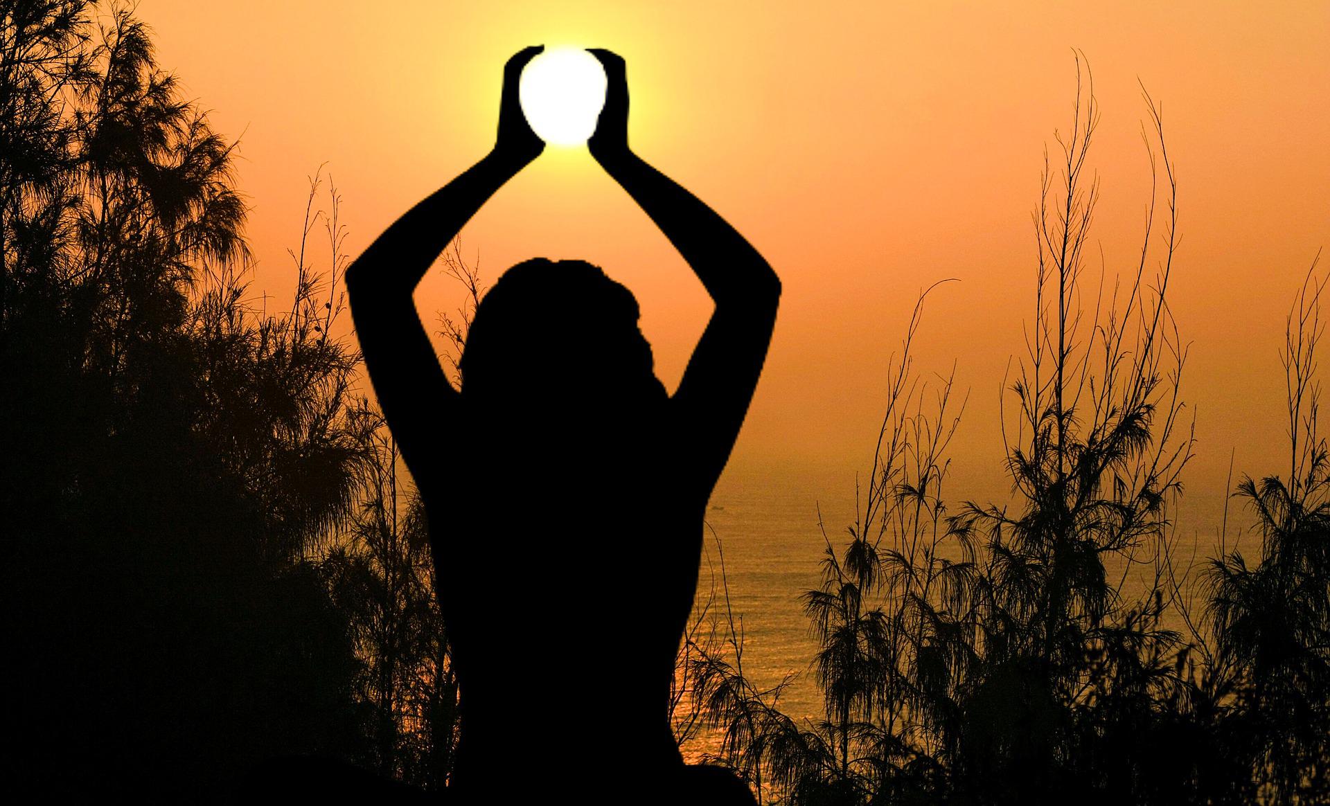 The silhouette of a woman with her arms raised, her hands appearing to be around the soft sun. We see her from the back. There are trees and grasses also in silhouette. The sea is visible.