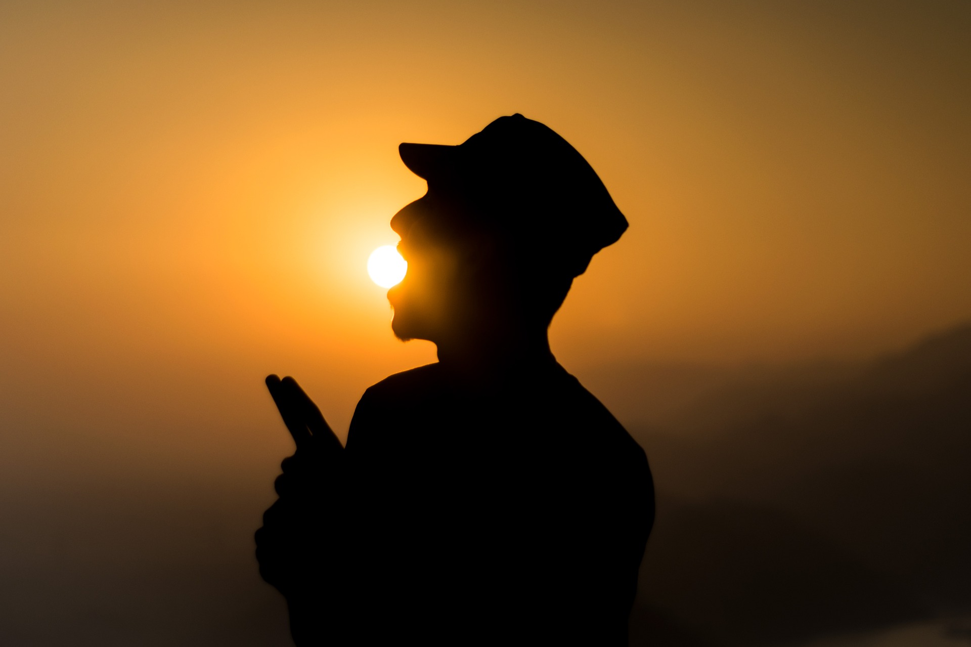 The silhouette of a standing man with a cap on appears to be literally eating the sun. The sun is low on the horizon, above some mountains.