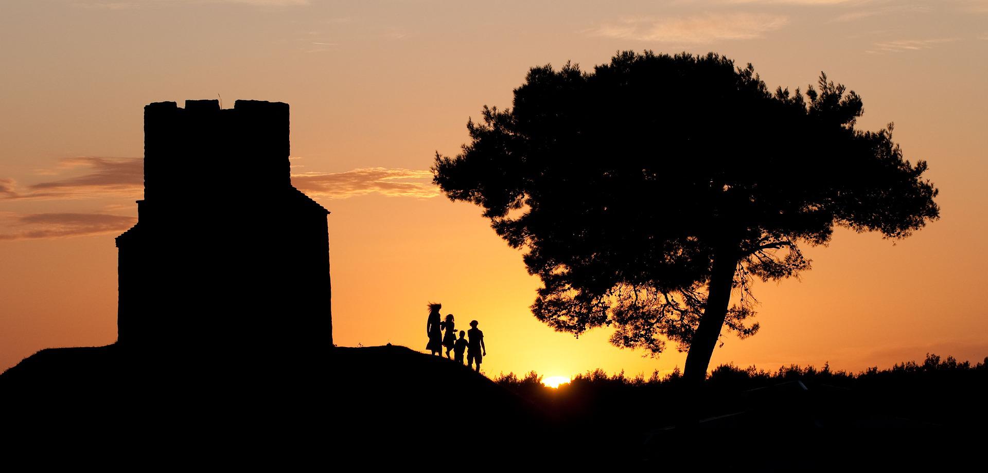 We see the silhouette of a family, a tree and a small home with a castellated roof sited on a hillock. The family watches the sun on the horizon.