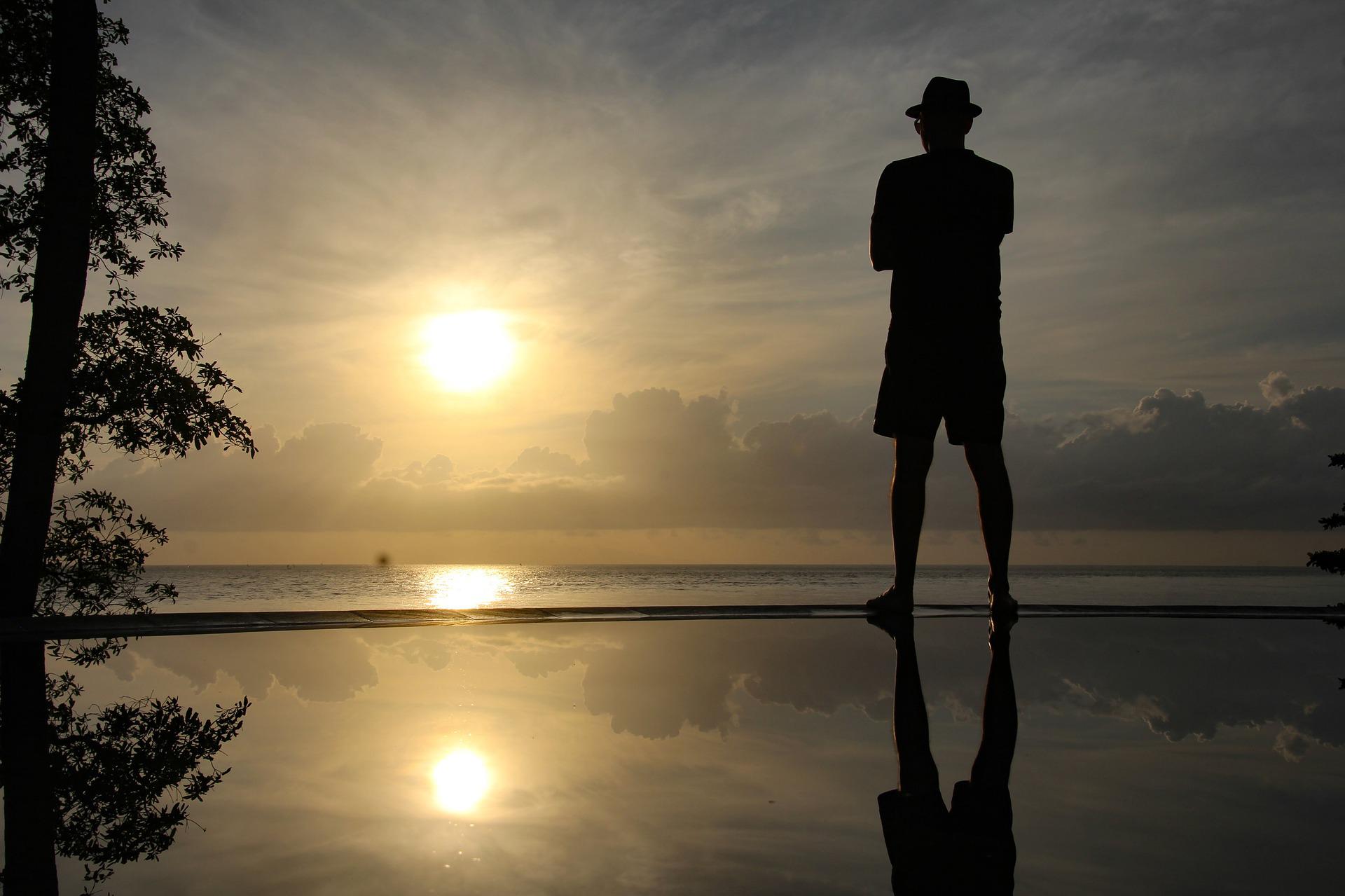 A man with a hat, spectacles and arms crossed stands gazing at the horizon sun. We see him from back. He stands on the edge of a zero edge swimming pool, looking quietly over the sea at the freeing sun. There is a large tree on the left.