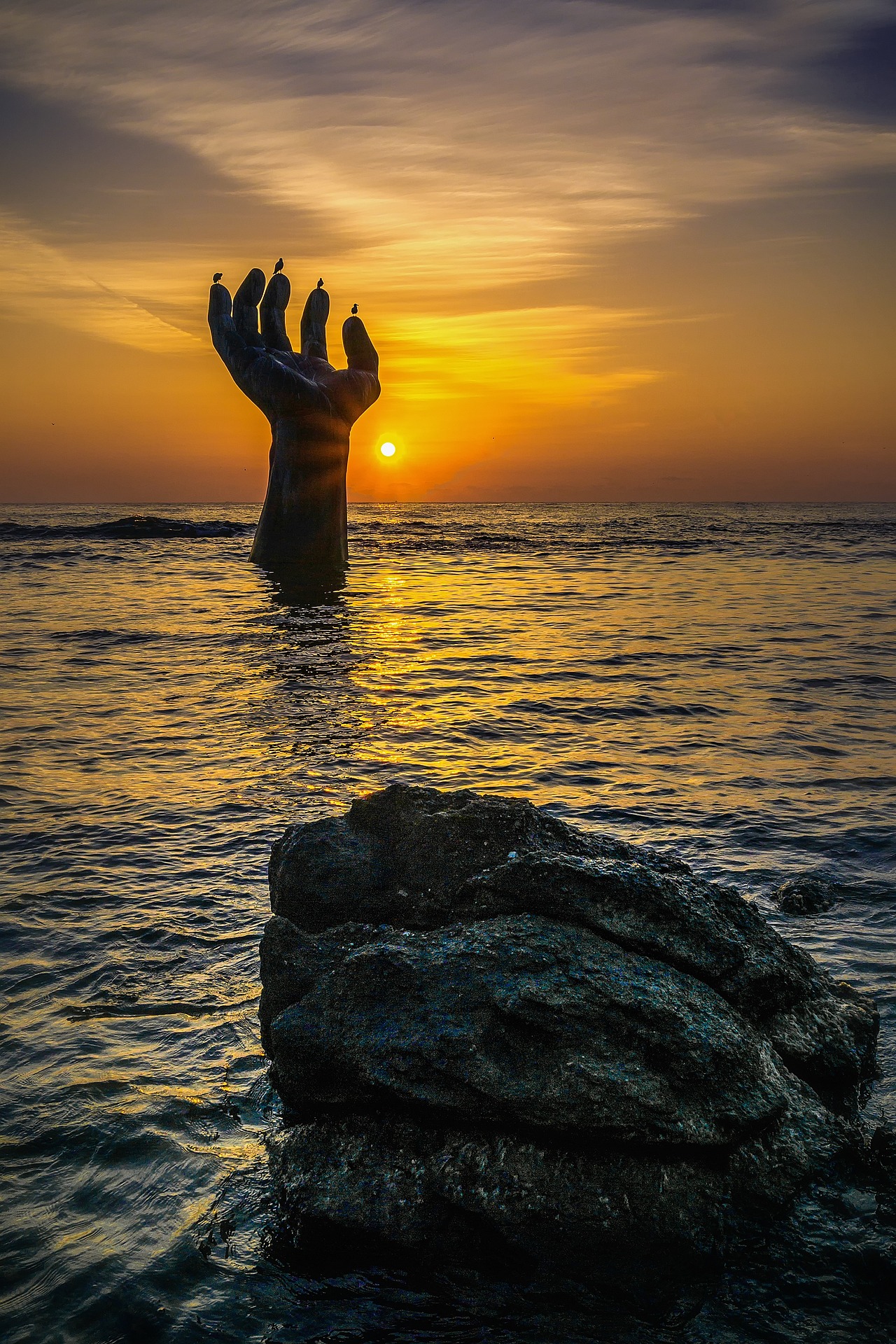 A photo of "The Hand of Coexistence", Homigot, Pohang, South Korea. A bronze hand sculpture sticks out of the sea. It is sunrise and the sun is visible too. The hand is upturned, open. Some seagulls perch on the tips of the fingers.