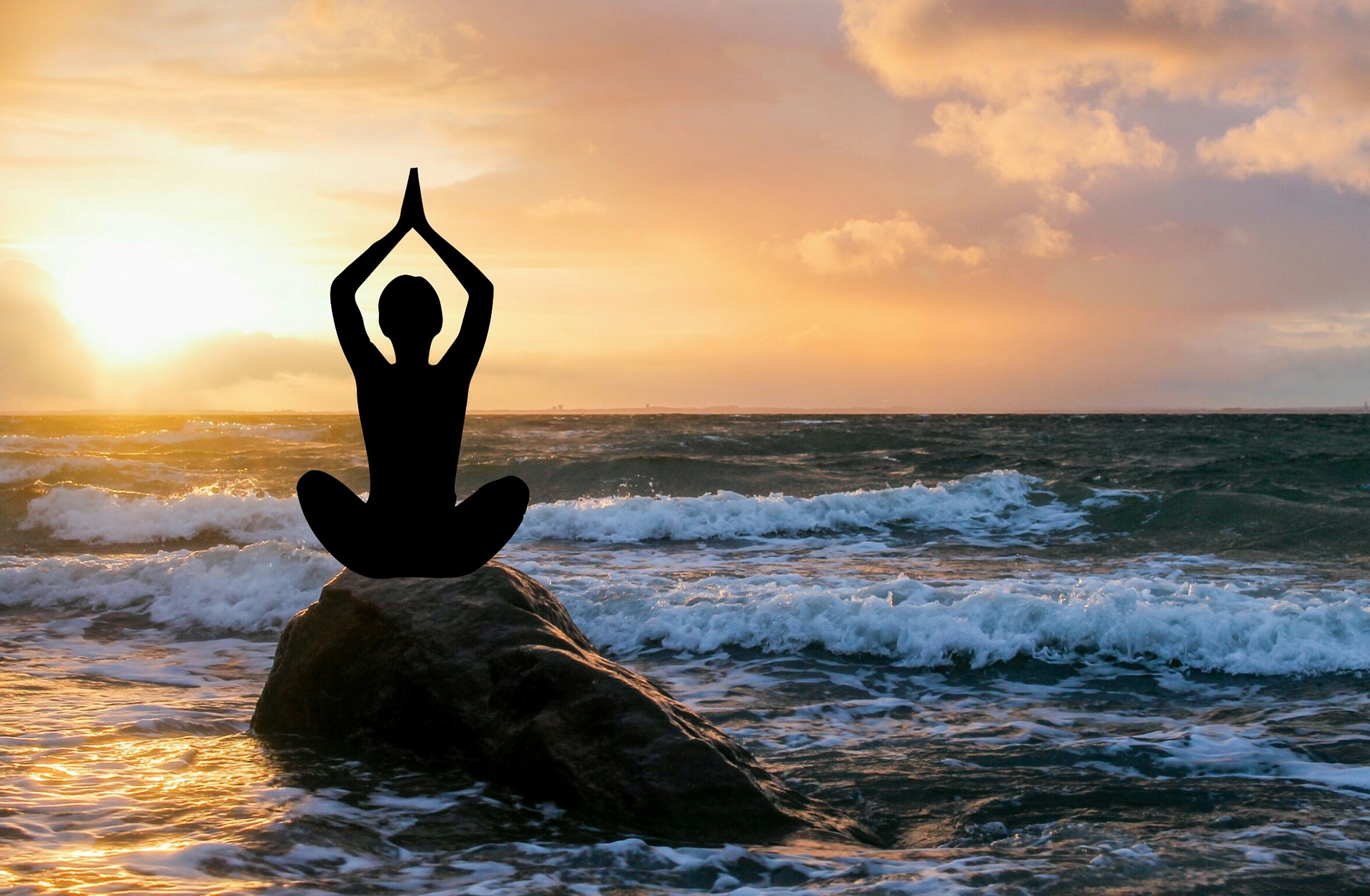 As the sun rises or sets, a human silhouette sits atop a large rock in the sea's surf. It is at a beach. The person is meditating and has their hands in a prayerful pose above their head. The blues - sea, surf, rock -of the lower part of the photo contrast with the warmer colours - sun, sky, clouds - of the upper photo.