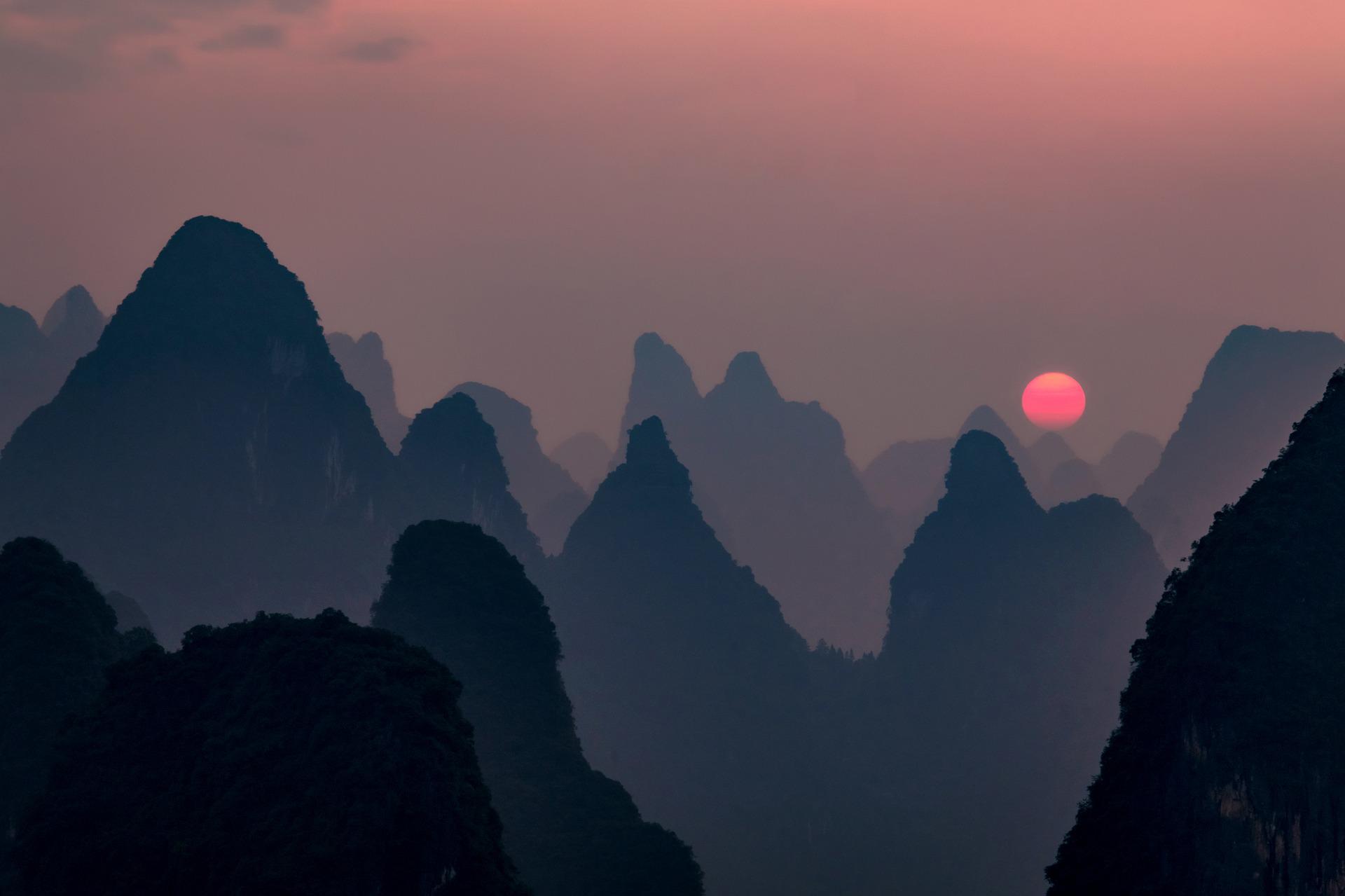 Amidst darkened Chinese hills, there is a rosy sky and rosy-red sun on the distant horizon. A beautiful mood.
