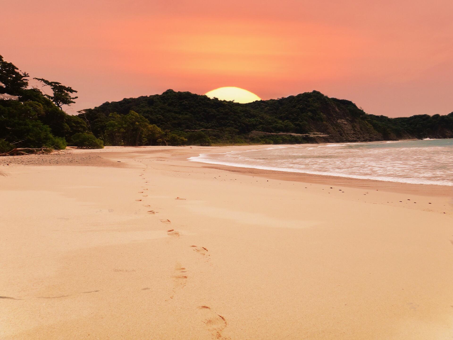 A sun is rising above a Costa Rica coast. A large sun is just beginning to emerge above a tropical jungle. The sky is a beautiful peach colour. In the foreground, beach and surf stretch towards the jungle. There are footprints in the sand.