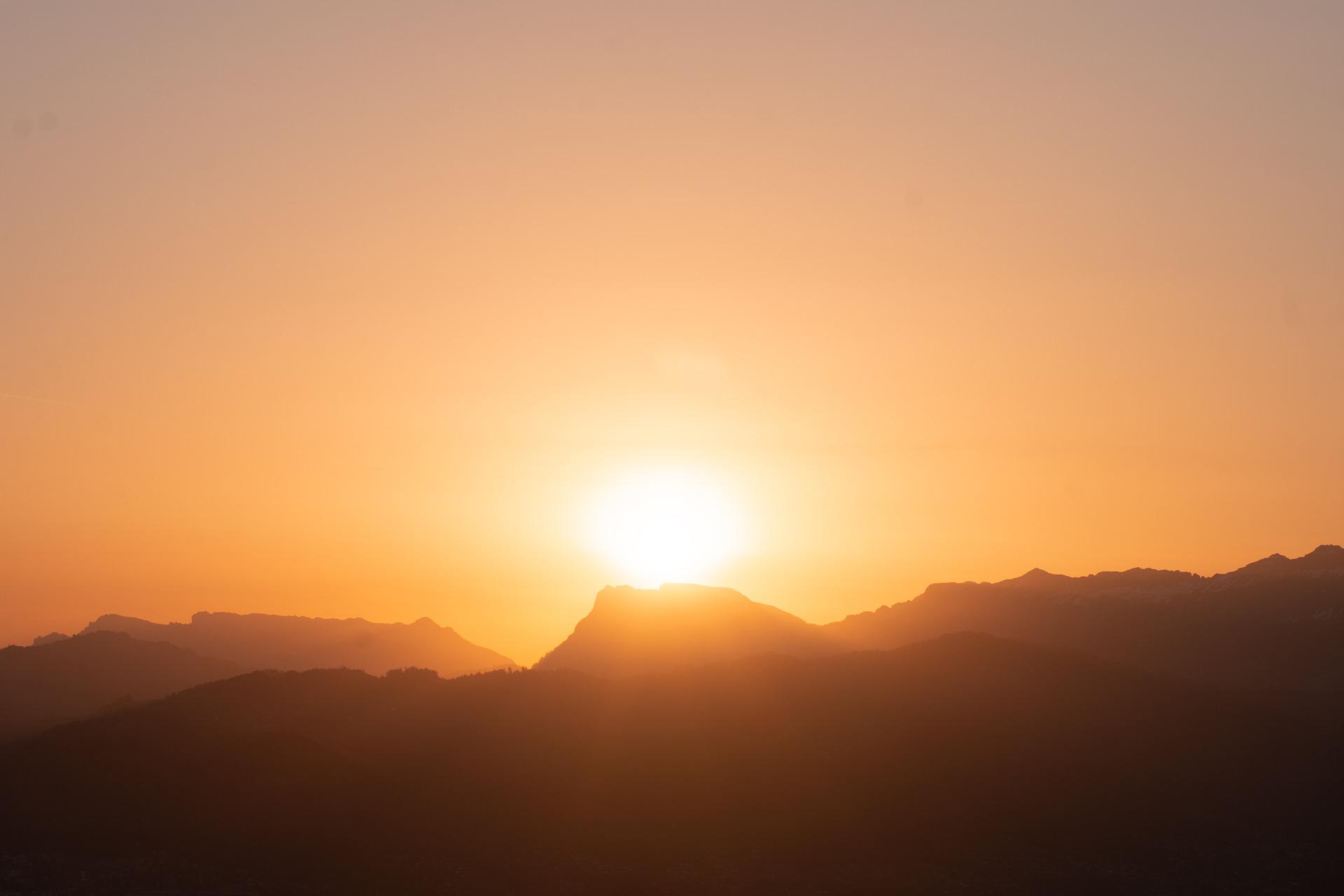 The sun is rising strongly over a mountain range. It appears momentarily to sit atop a mountain peak.