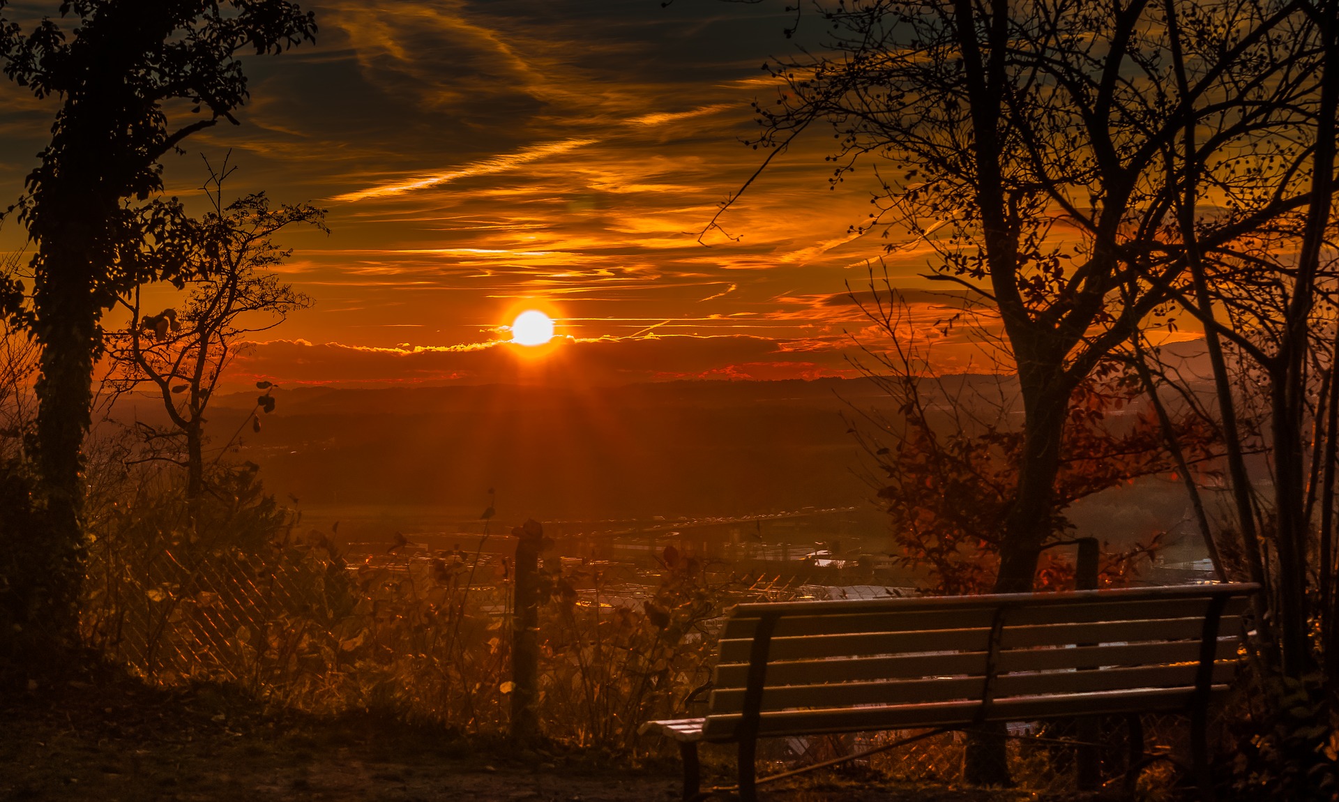 A sunrise is happening over a town. We are elevated, on a wooded hill overlooking the town. There is an empty bench if you want to be seated.