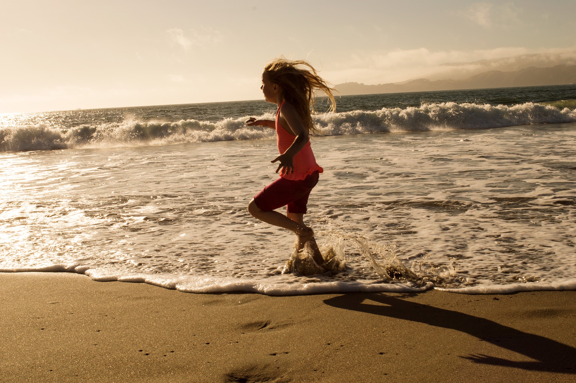 A girl on the beach is running on the incoming surf, her knees lift high, her arms/hands are outstretched