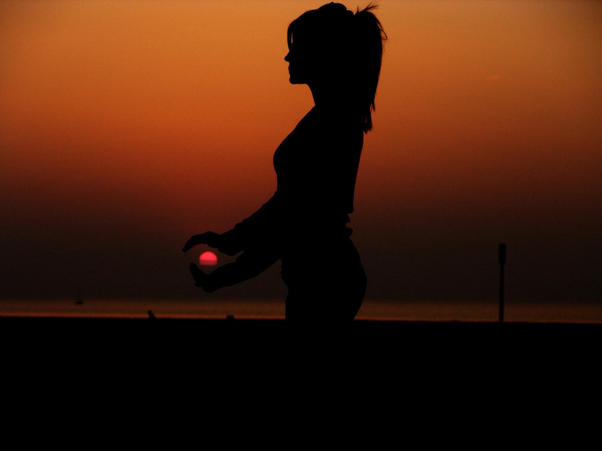 In the foreground, the silhouette of a standing meditative woman by the sea. Her hands appear to be around the red sun on the horizon, as if holding it in her hands.