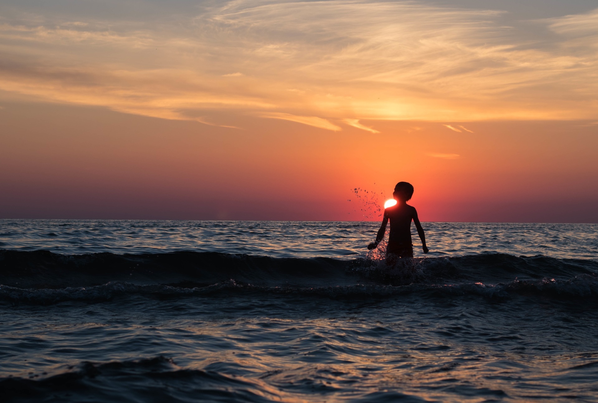 The sun rises on the horizon and the silhouette of a boy is wading in the sea