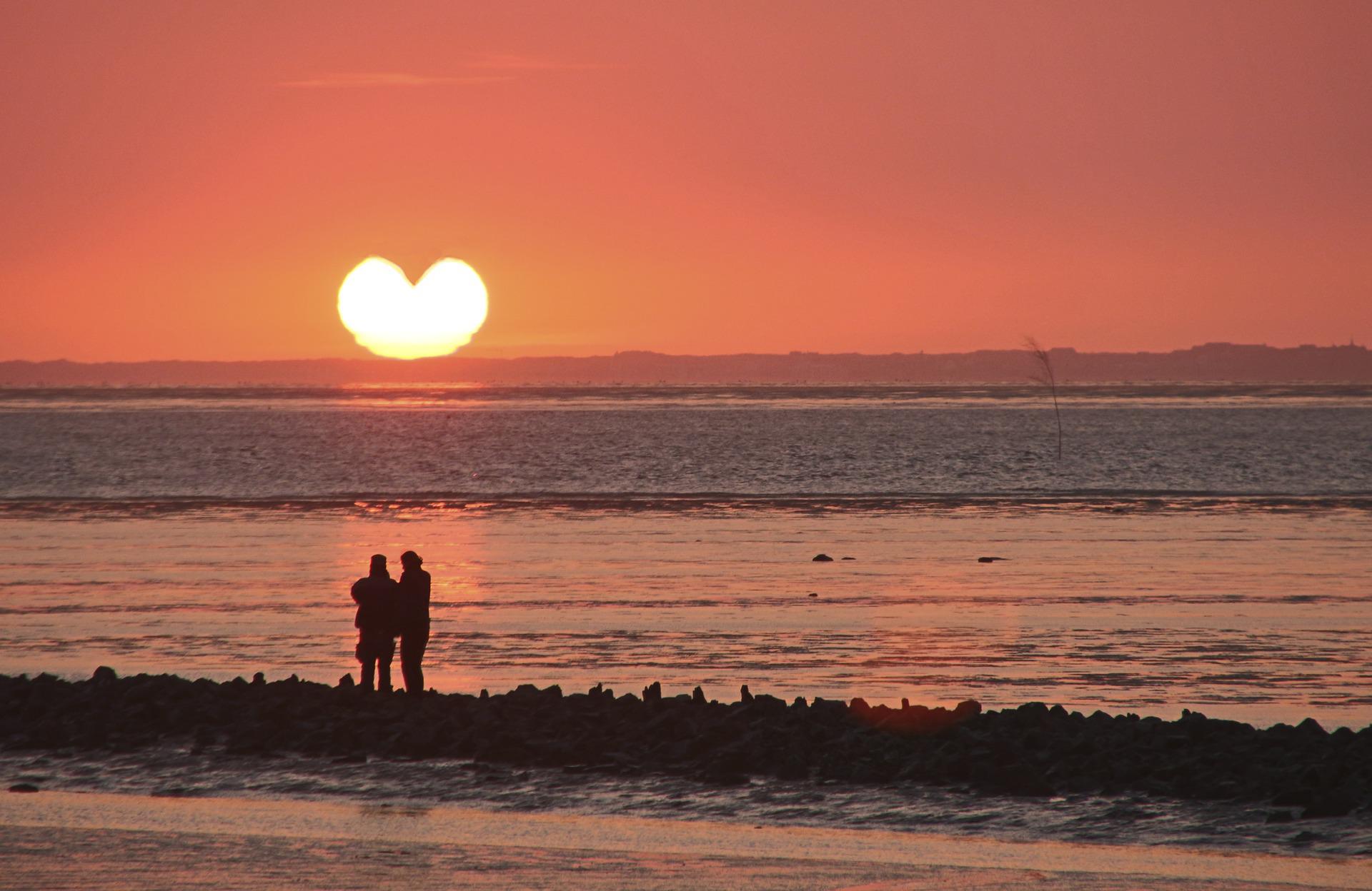 A couple on a beach gaze at the sun, on the horizon. The waves lap in. The sun is heart-shaped, almost certainly due to digital manipulation (not a fortunate cloud formation). The sky is a very warm, soft pink-orange.