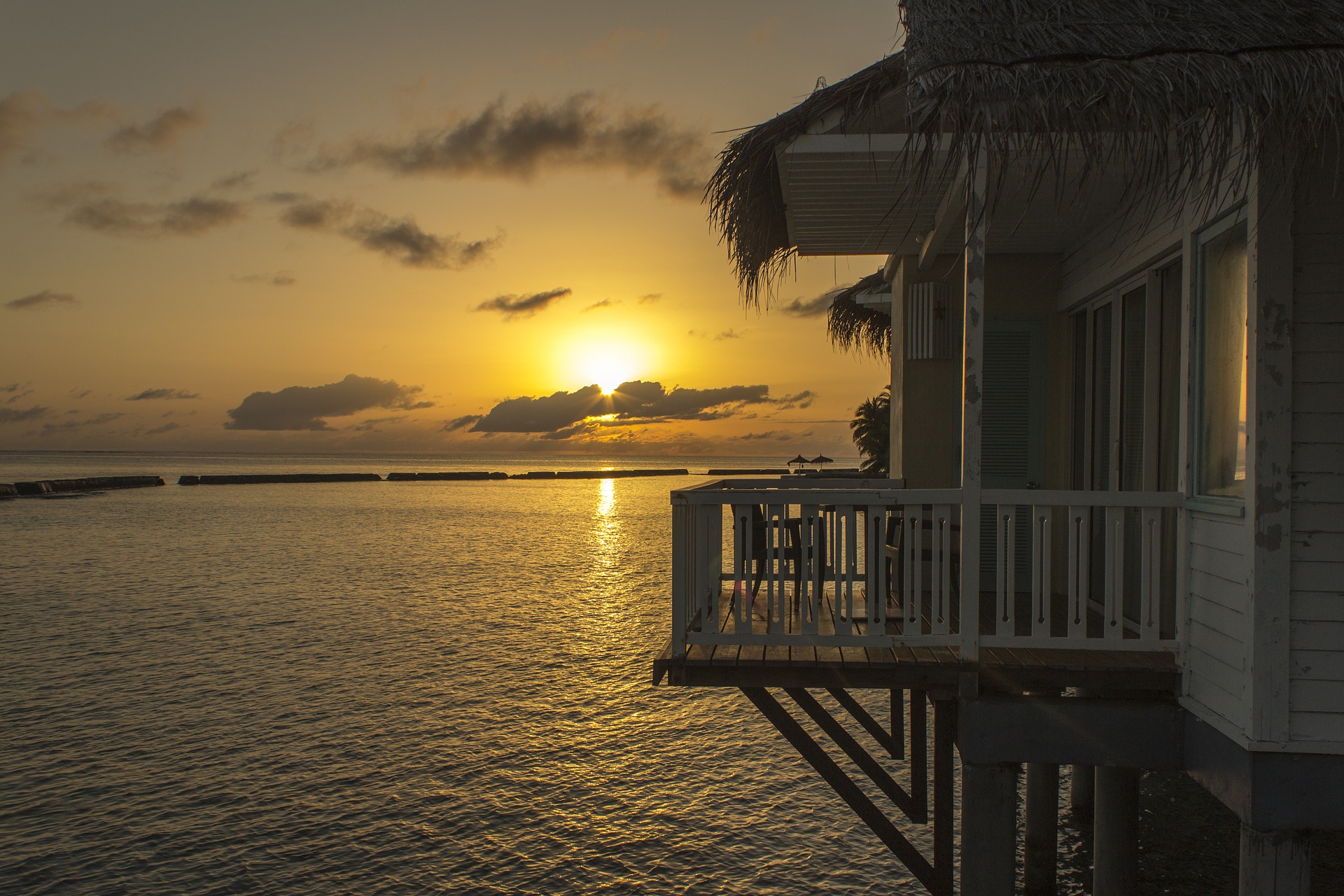 We see a bungalow hut with thatch roof. It is on stilts in the sea, in the Maldives. The sun is rising.