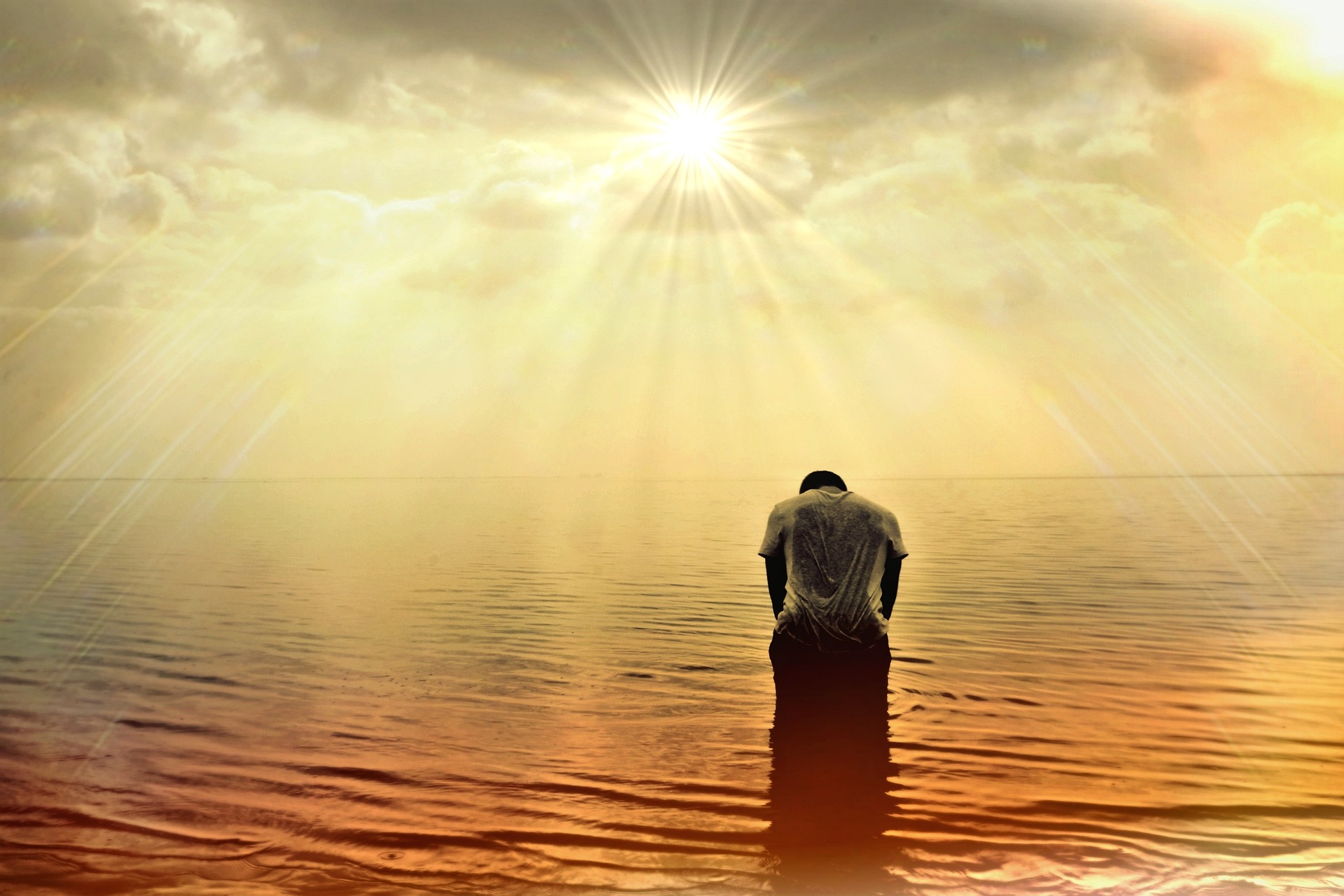 A man is kneeling in a shallow sea, bowing to the spectacular rising sun