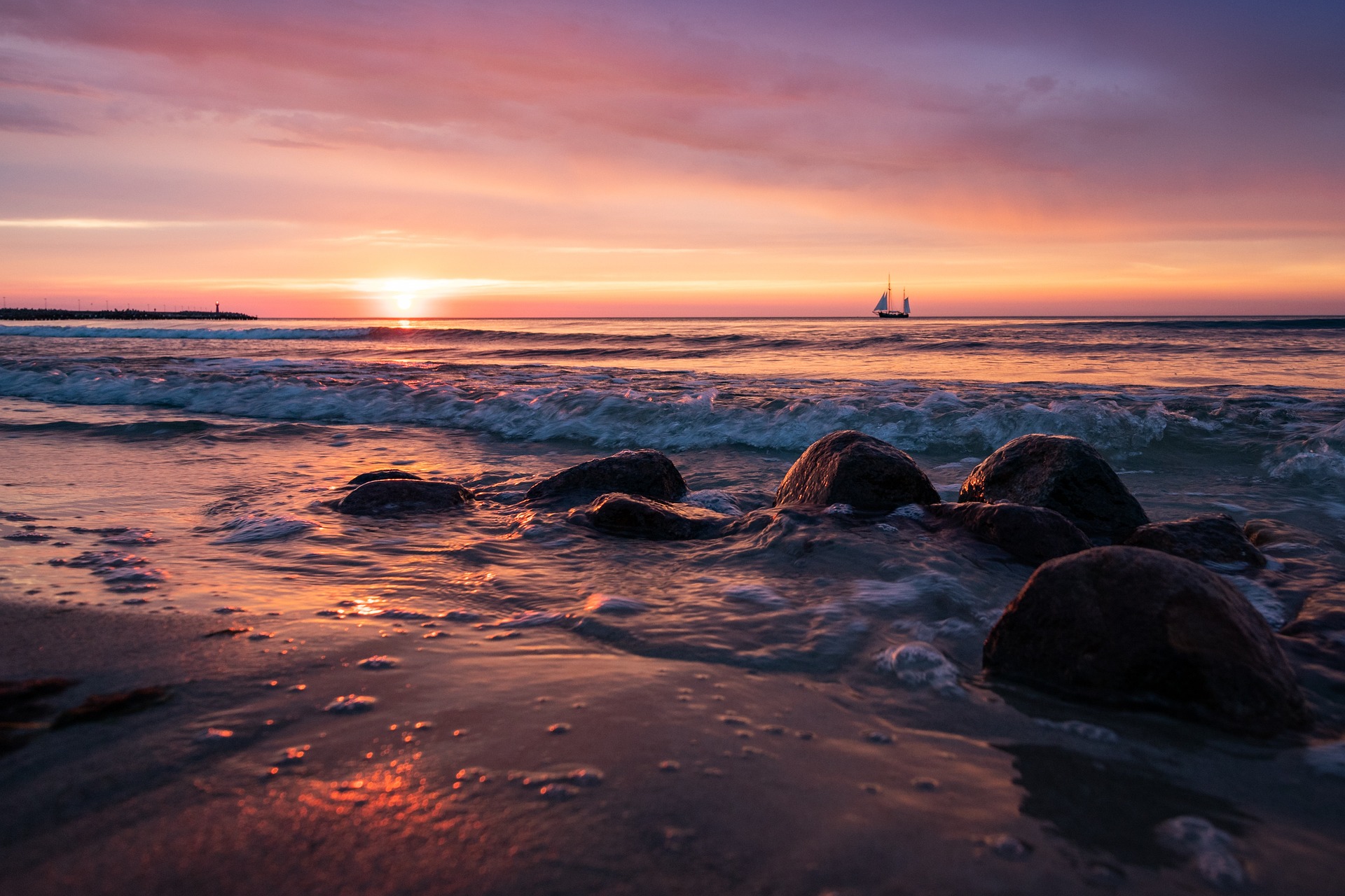 A sunrise over a Baltic Sea beach. A large sailing ship is on the horizon. Some large rocks are in the foreground.