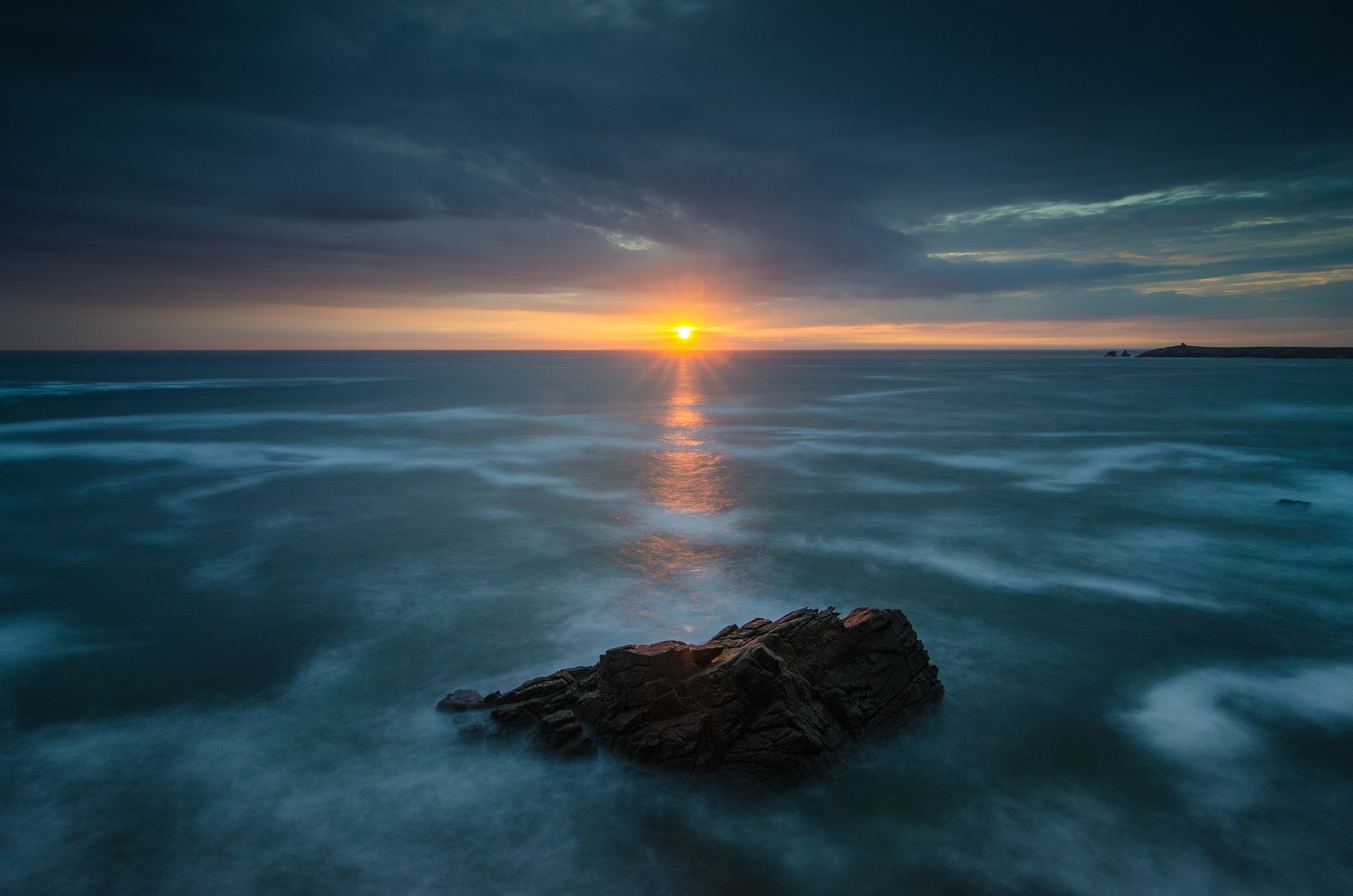 A sunrise over the ocean, seen from France. A large rock is in the foreground.