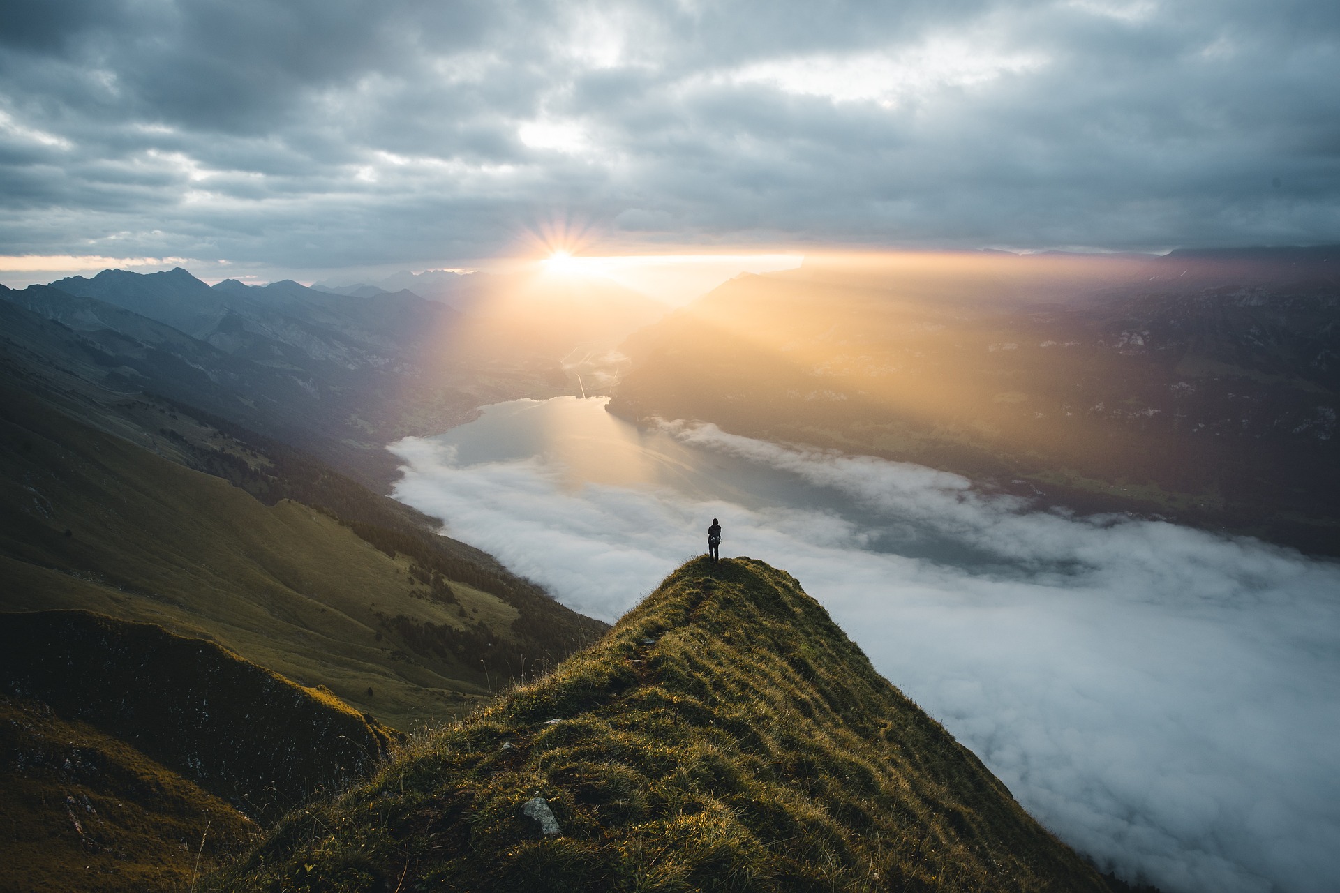 A sunrise over a lake viewed from atop a mountain. In the foreground, a person stands quietly looking on from a slightly lower peak. Mist sweeps across the lake.
