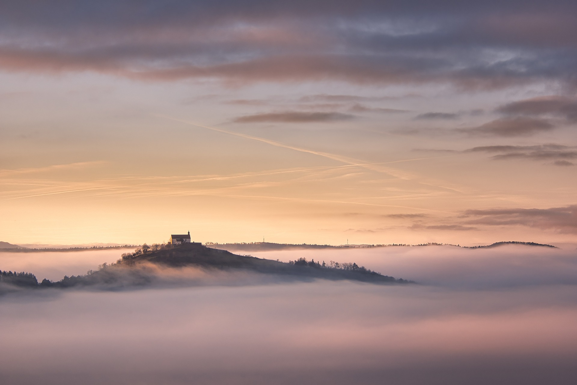 A sunrise somewhere in the world. A hill with a house on top of it juts out from a misty landscape.