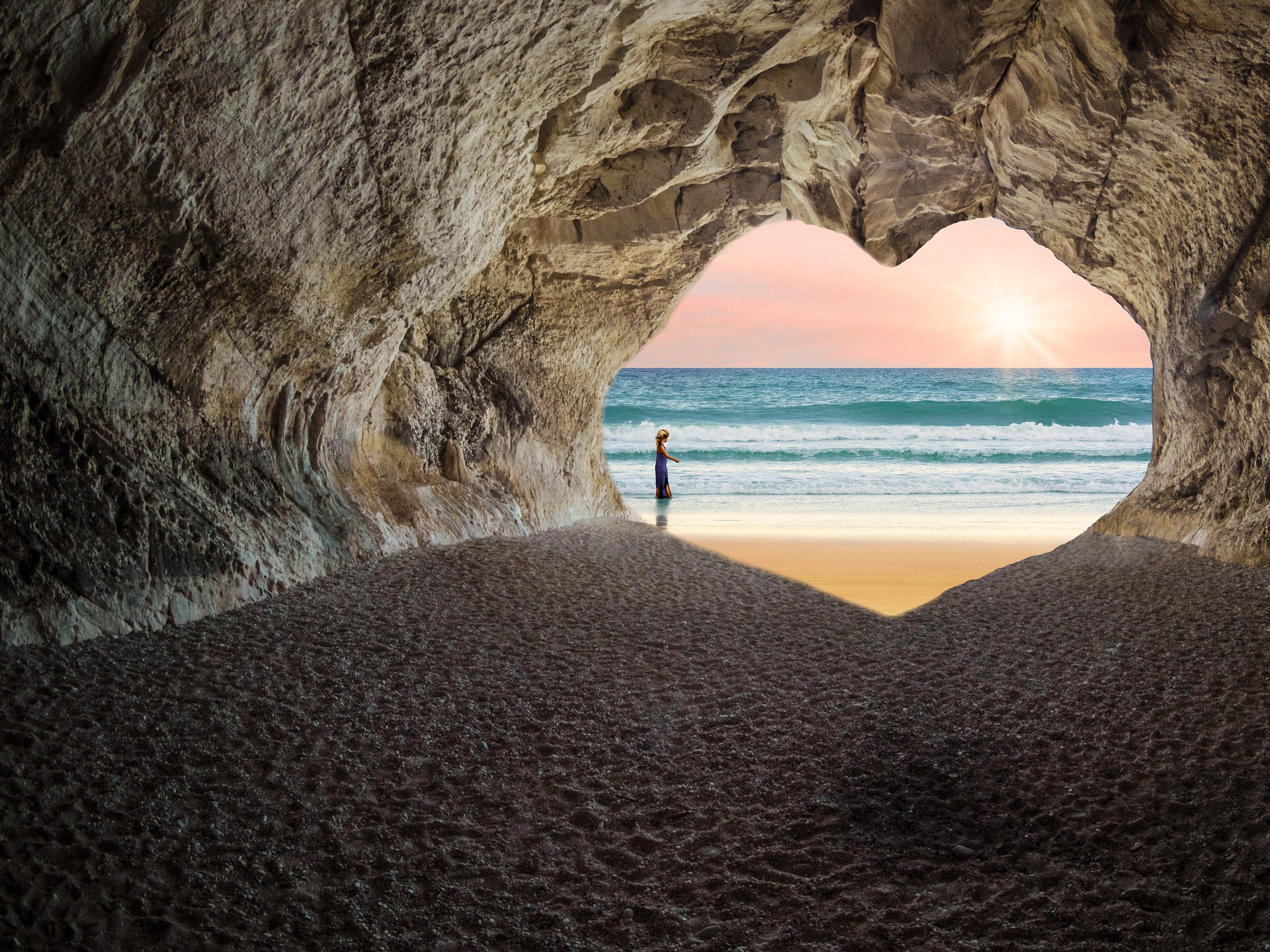 We are looking out from the inside of a beach cave. Through its beautiful heart shaped entrance, we can soft blue waves gently lapping onto the sand. The soft sun is on the horizon lighting everything. A person is calmly standing where the surf meets the sand.