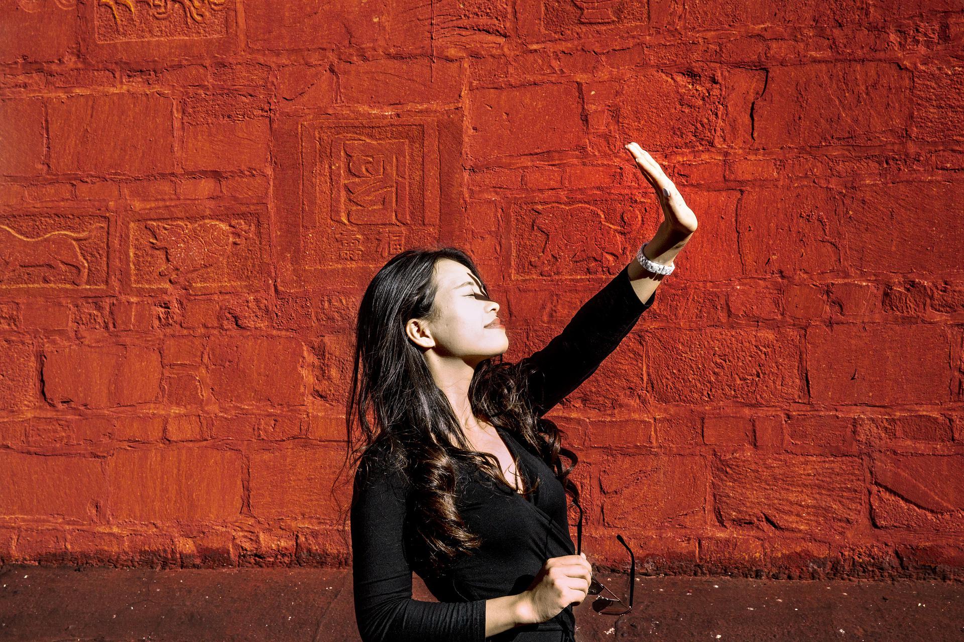 A young Asian woman is standing in front of a red Buddhist wall in Tibet. She has her eyes closed and gazes up at the sun. One hand is raised as she seems to adjust herself to sunning.
