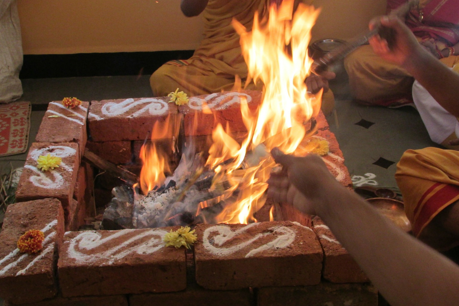 We see a square altar of bricks, a few bricks high, with ritual sand symbols and flowers on top of the bricks. A fire is lit within the altar and people are adding offerings.